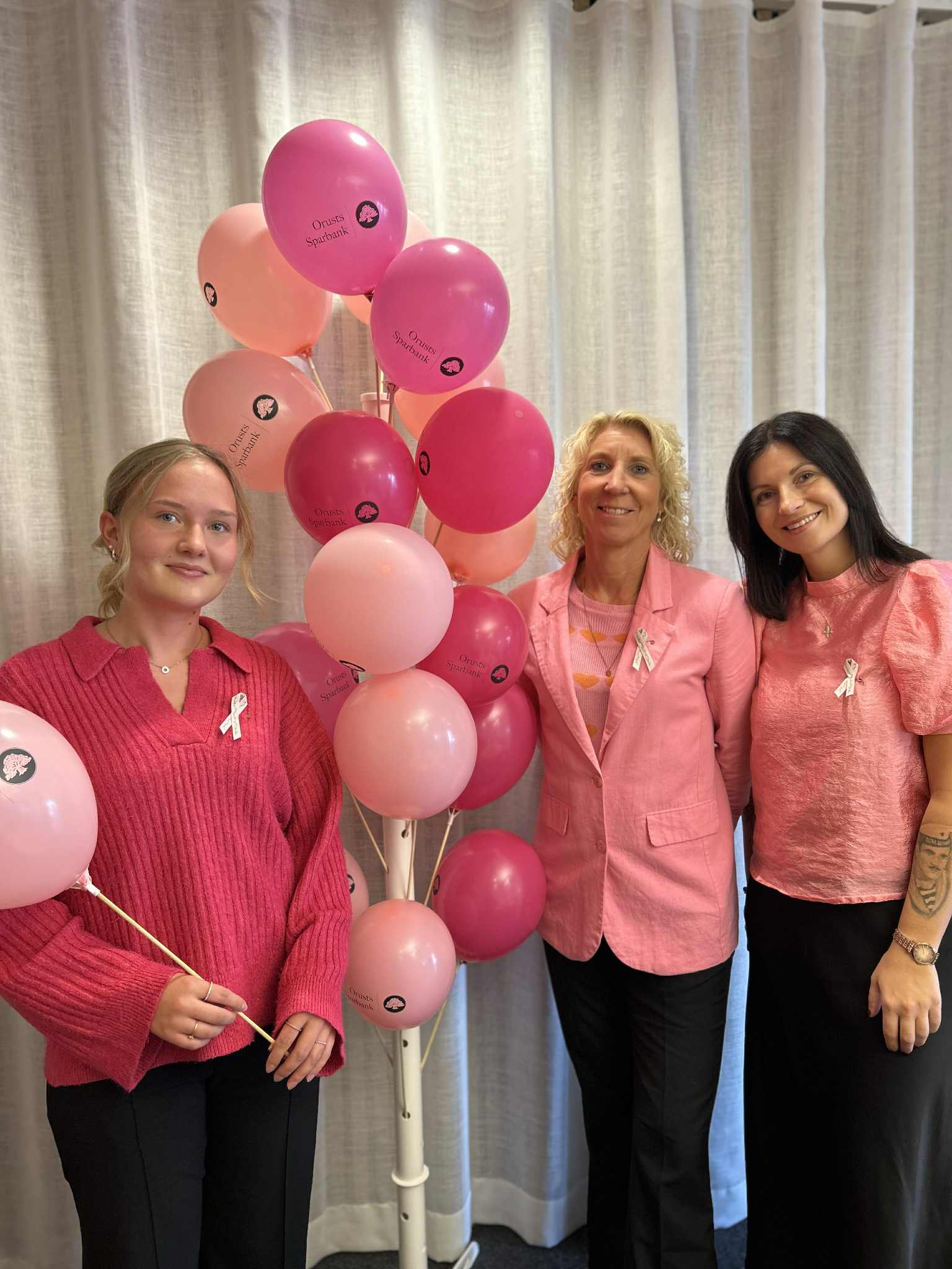 Tre kvinnor i rosa kläder står bredvid rosa ballonger med Orusts Sparbanks logotyp, till stöd för Rosa Bandet.
