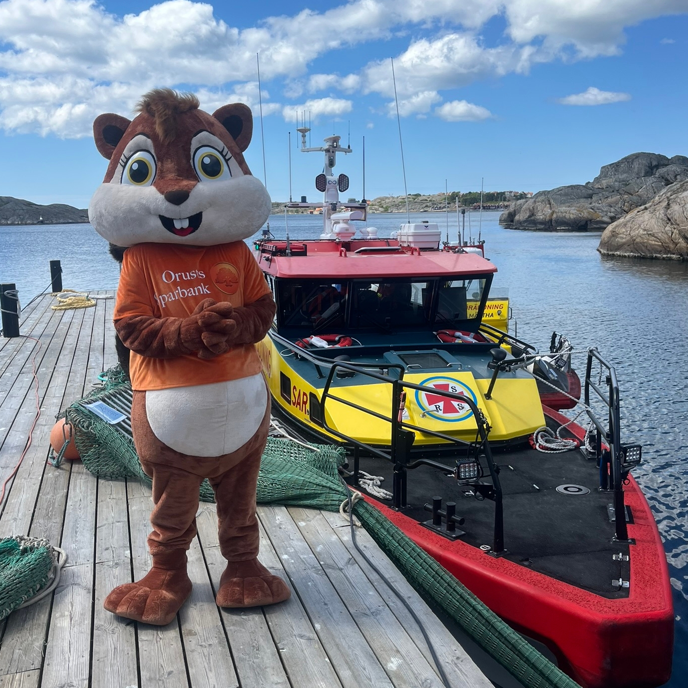 Orusts Sparbanks ekorre-maskot Ella står på en brygga bredvid en rödgul sjöräddningsbåt under blå himmel.