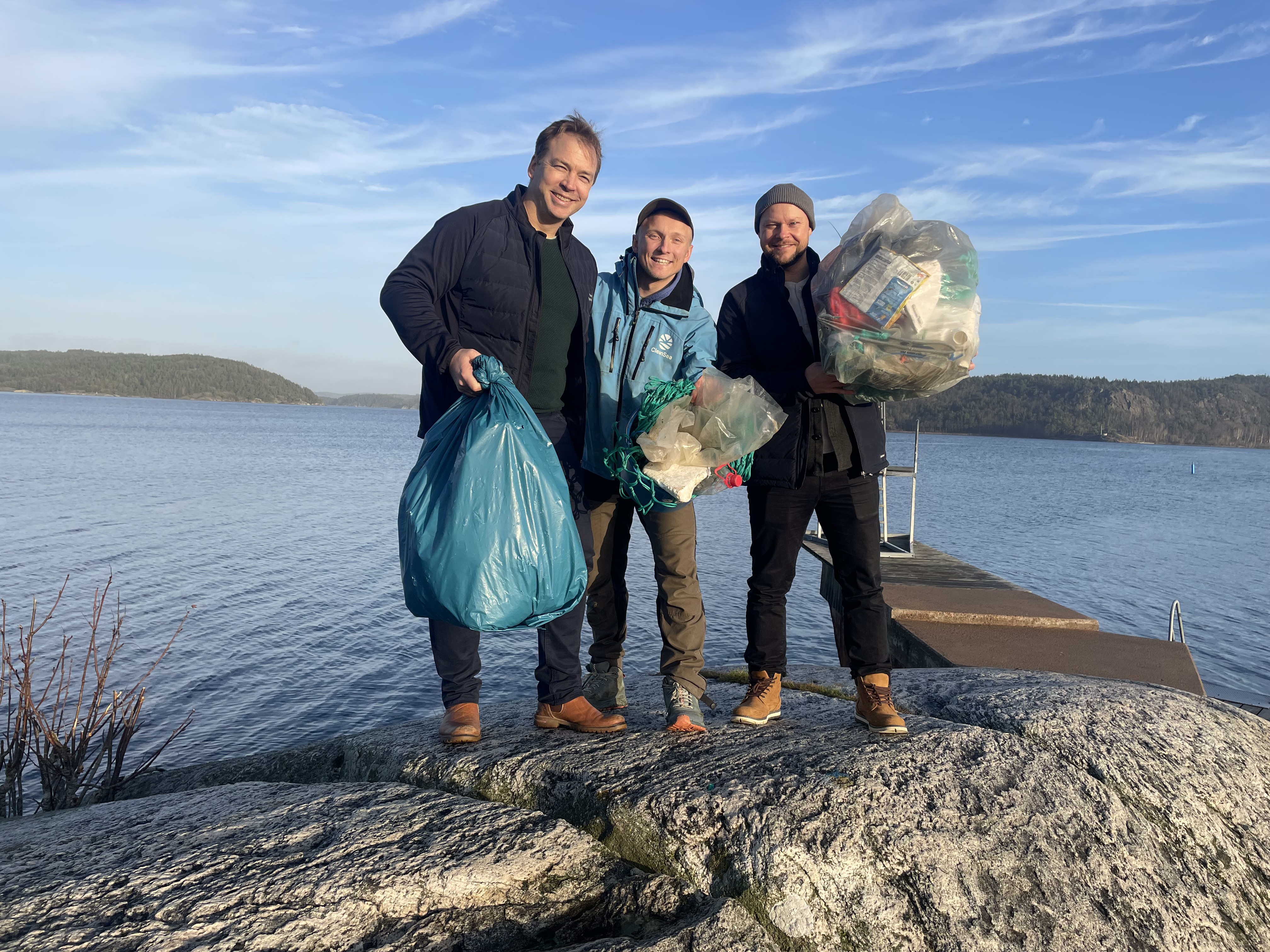 Tre personer står vid vattnet och håller upp påsar med insamlat skräp efter strandstädning.