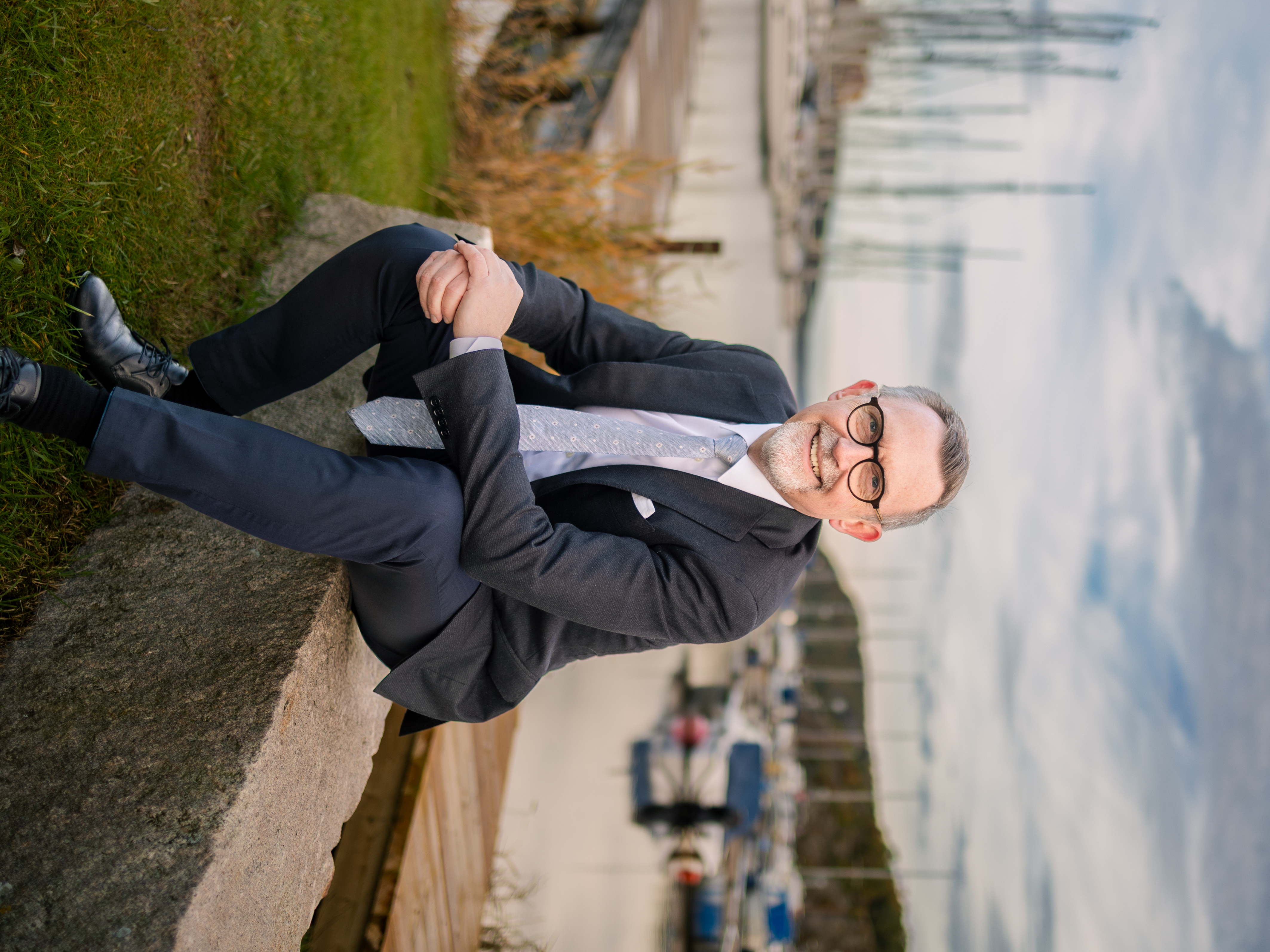 Leende man i kostym sitter vid en småbåtshamn med segelbåtar i bakgrunden.
